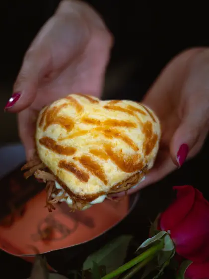 Mãos segurando um pão de queijo recheado em formato de coração, com rosas